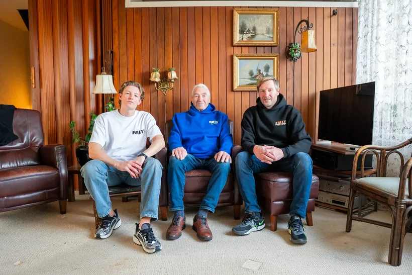 Drie mannen van verschillende generaties in kleding van FMLY op een bank in een huiskamer met houten wanden