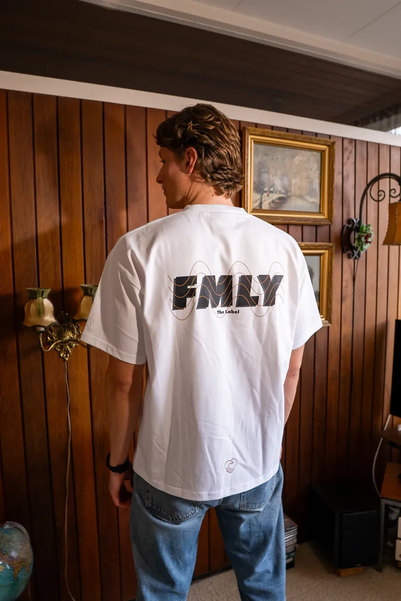 Man van achteren in een wit T-shirt met het FMLY-logo op de rug, tegen een houten wand