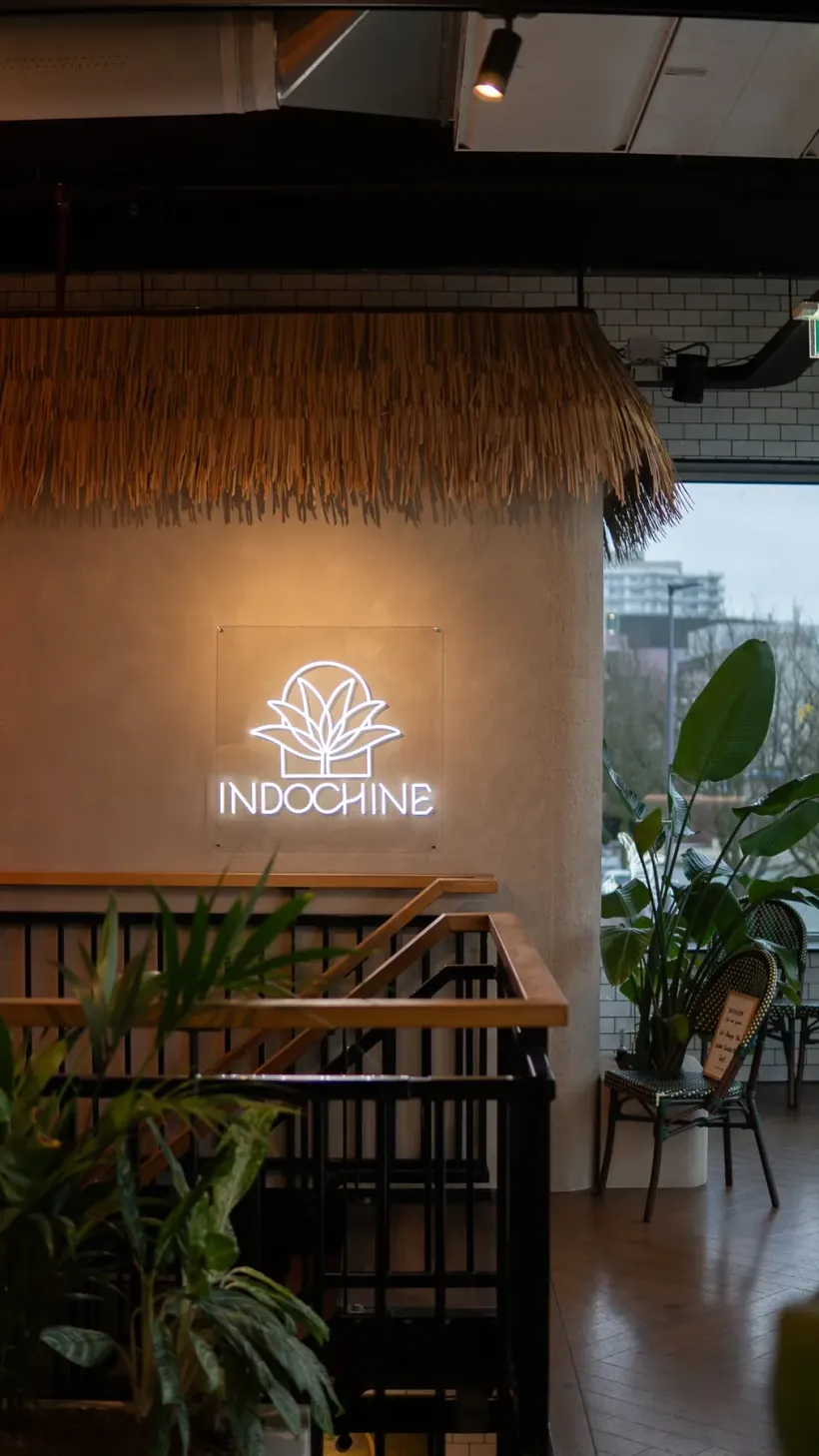 Lichtgevend logo van Indochine aan een wand met rieten plafond en planten bij de entree