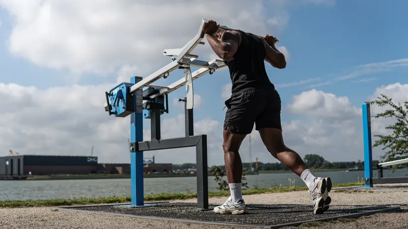 Sporter in zwarte sportkleding traint aan een blauw fitnesstoestel aan het water