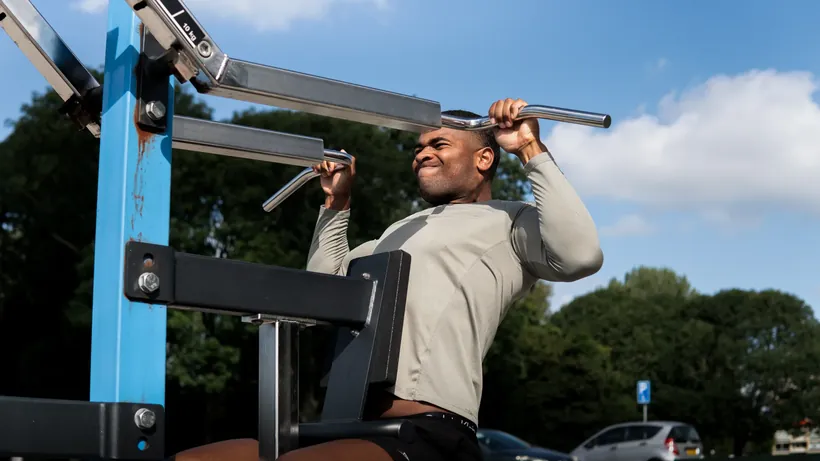 Sporter in een grijs shirt aan een outdoor fitnesstoestel onder een blauwe lucht