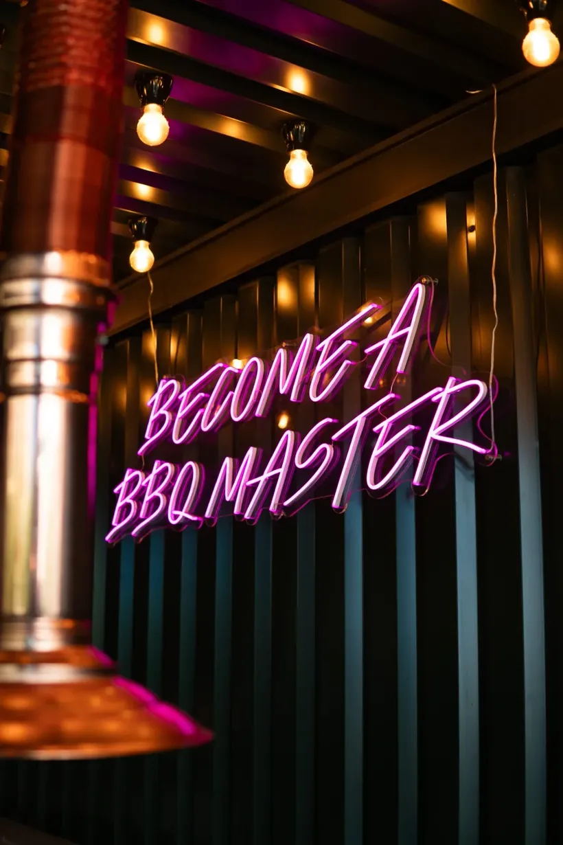 Roze neontekst Become a BBQ Master naast een koperen afzuigkap boven een tafel